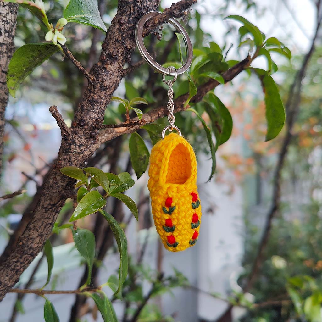 Yellow crocheted clog keyring with red and green accents hanging on a tree branch with green leaves
