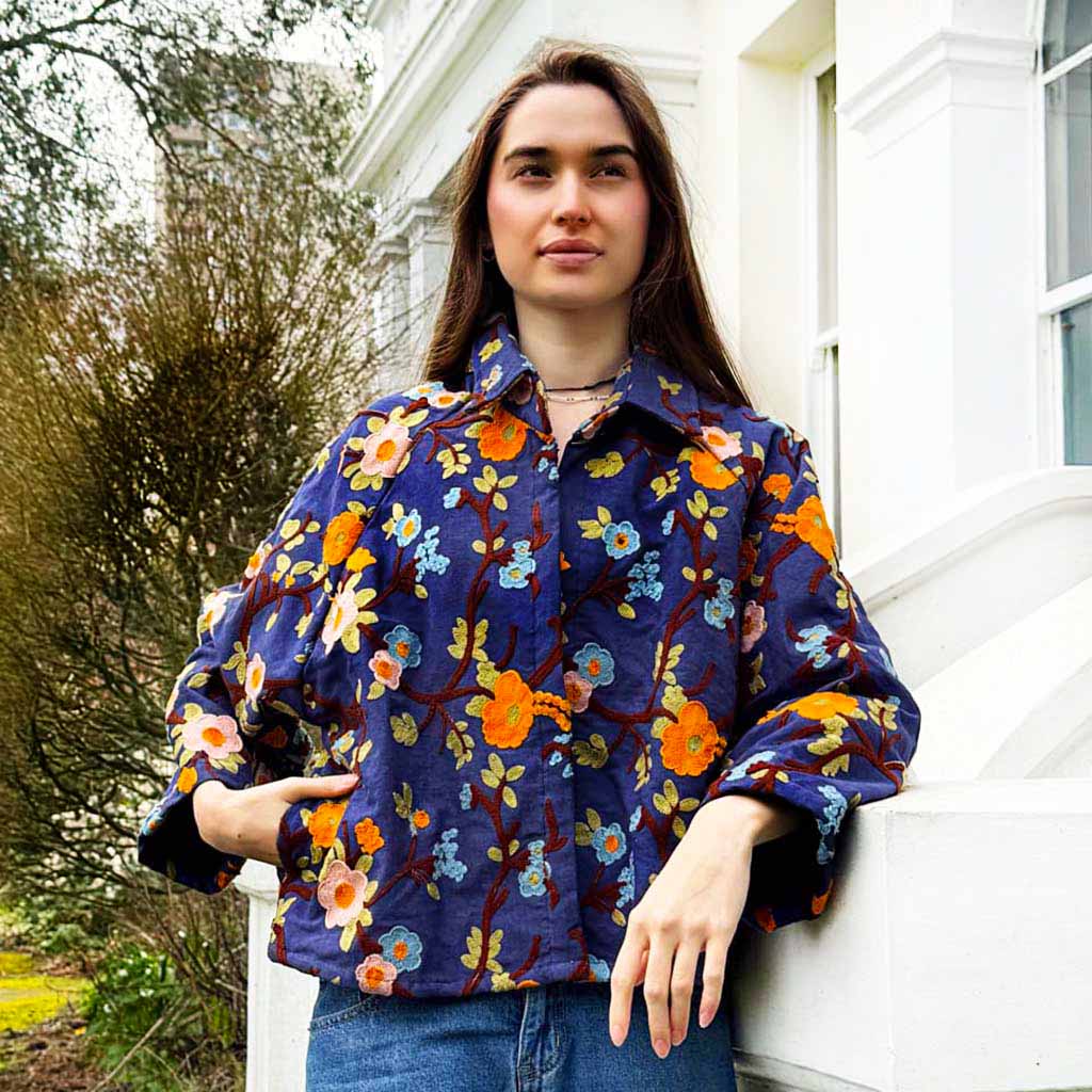 Model wearing a blue denim floral-patterned embroidered jacket on a next to a white building