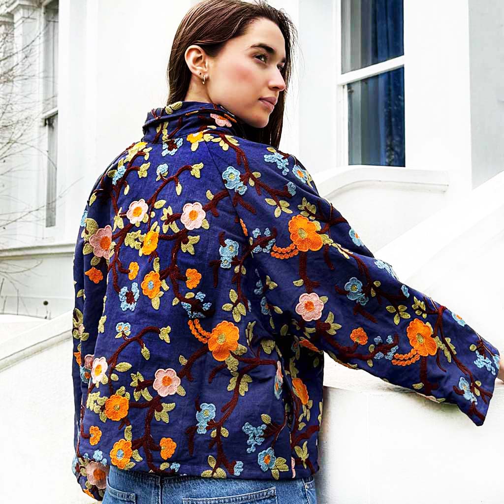 Model wearing a blue denim floral-patterned embroidered jacket on a next to a white building