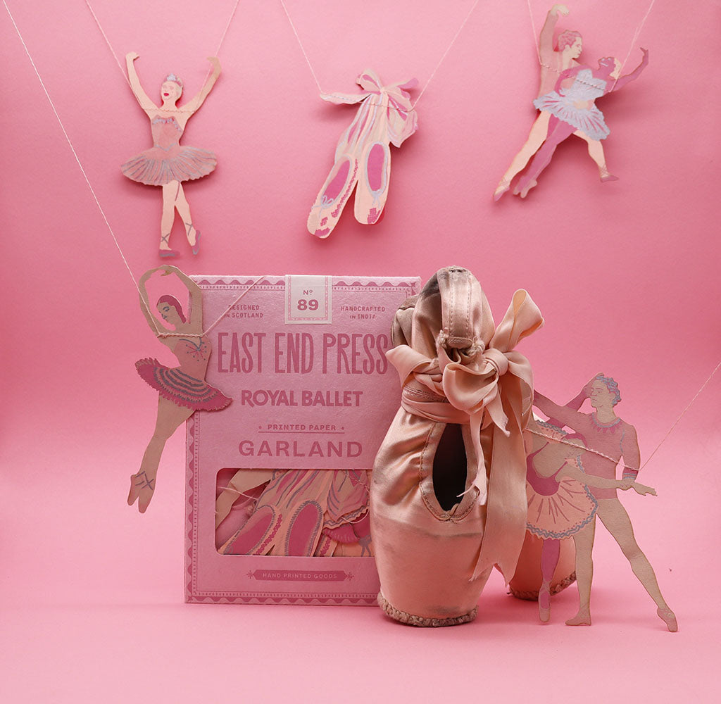 Ballerina-themed garland by East End Press, featuring a pink paper packaging labeled 'Royal Ballet' and 'Garland' alongside multiple pink and beige paper ballerina and pointe  shoe cutouts arranged against a solid pink background.