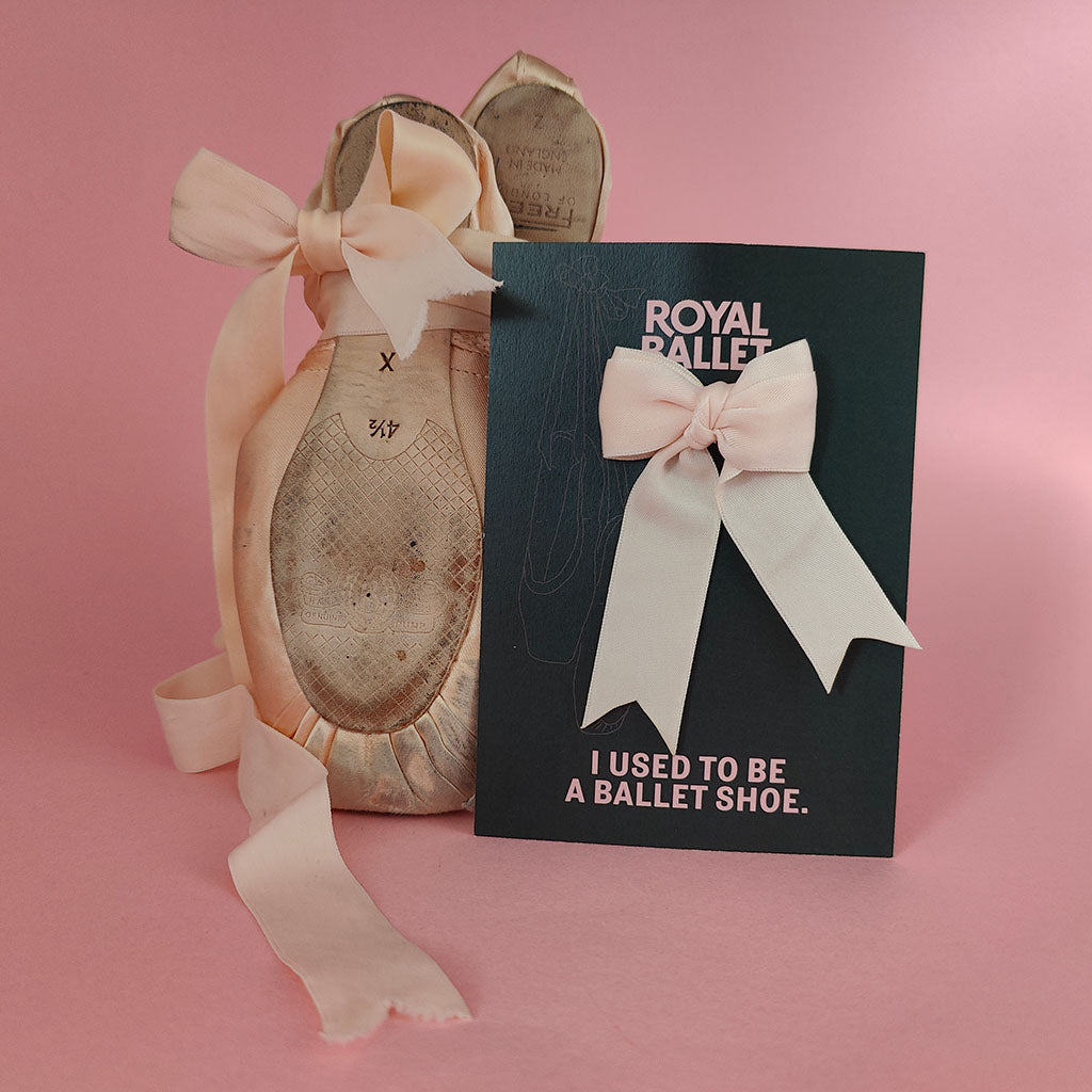 Pink satin ballet pointe shoes with a long ribbon stands next to a black card that reads 'ROYAL BALLET - I USED TO BE A BALLET SHOE' with a pink bow ribbon brooch attached, with a pink background