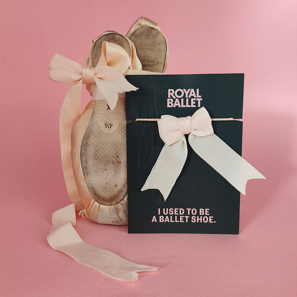 used pointe shoes on display with a recycled Encore! Hair Tie featuring recycled pointe shoe ribbon and elastic, the background is pink