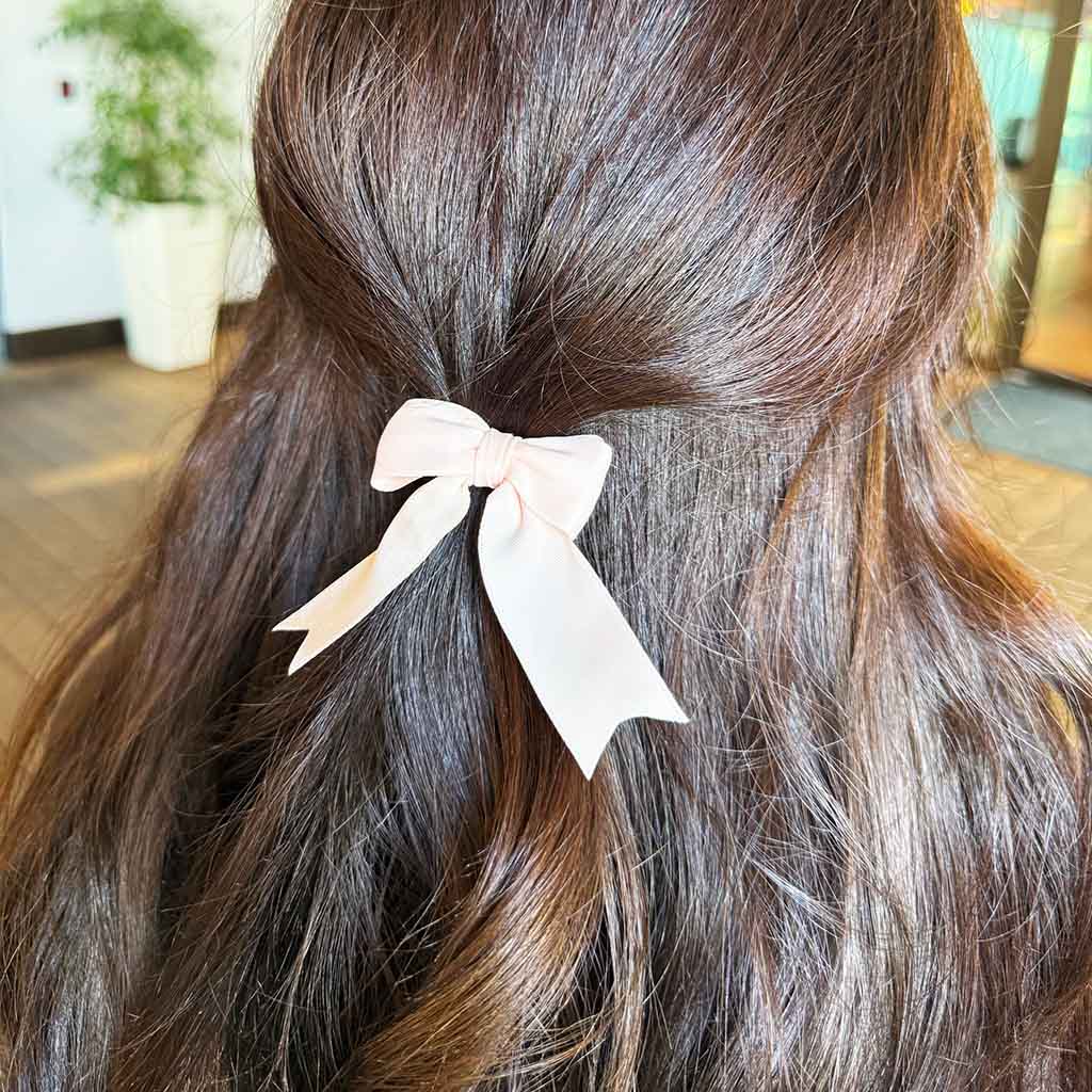 A close-up of a person's long, wavy brown hair with a pale pink satin bow hair tie secured in the middle.