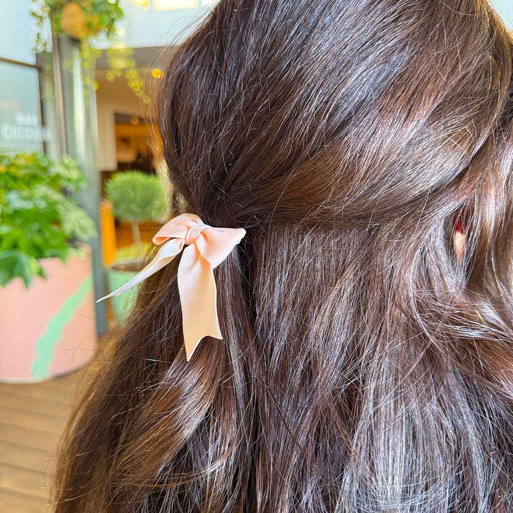 A light pink beige hair tie featuring a satin ribbon bow with two ribbon tails, shown on a plain white background made from recycled pointe shoes.