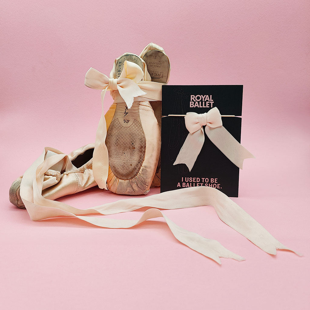 used pointe shoes on display with a recycled Encore! Hair Tie featuring recycled pointe shoe ribbon and elastic, the background is pink
