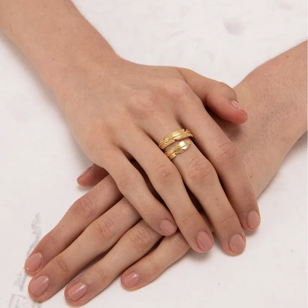 A feather ring worn on a person's hand, shown against a light neutral background. The hand is folded over with the fingers of the other hand resting on top.