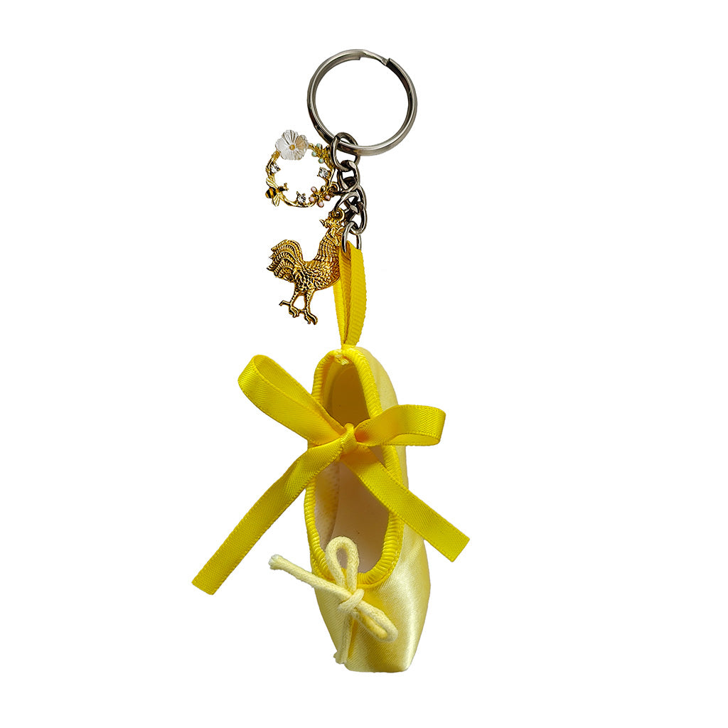 Yellow ballet shoe keychain with a ribbon and two charms a floral wreath and a chickenon a white background
