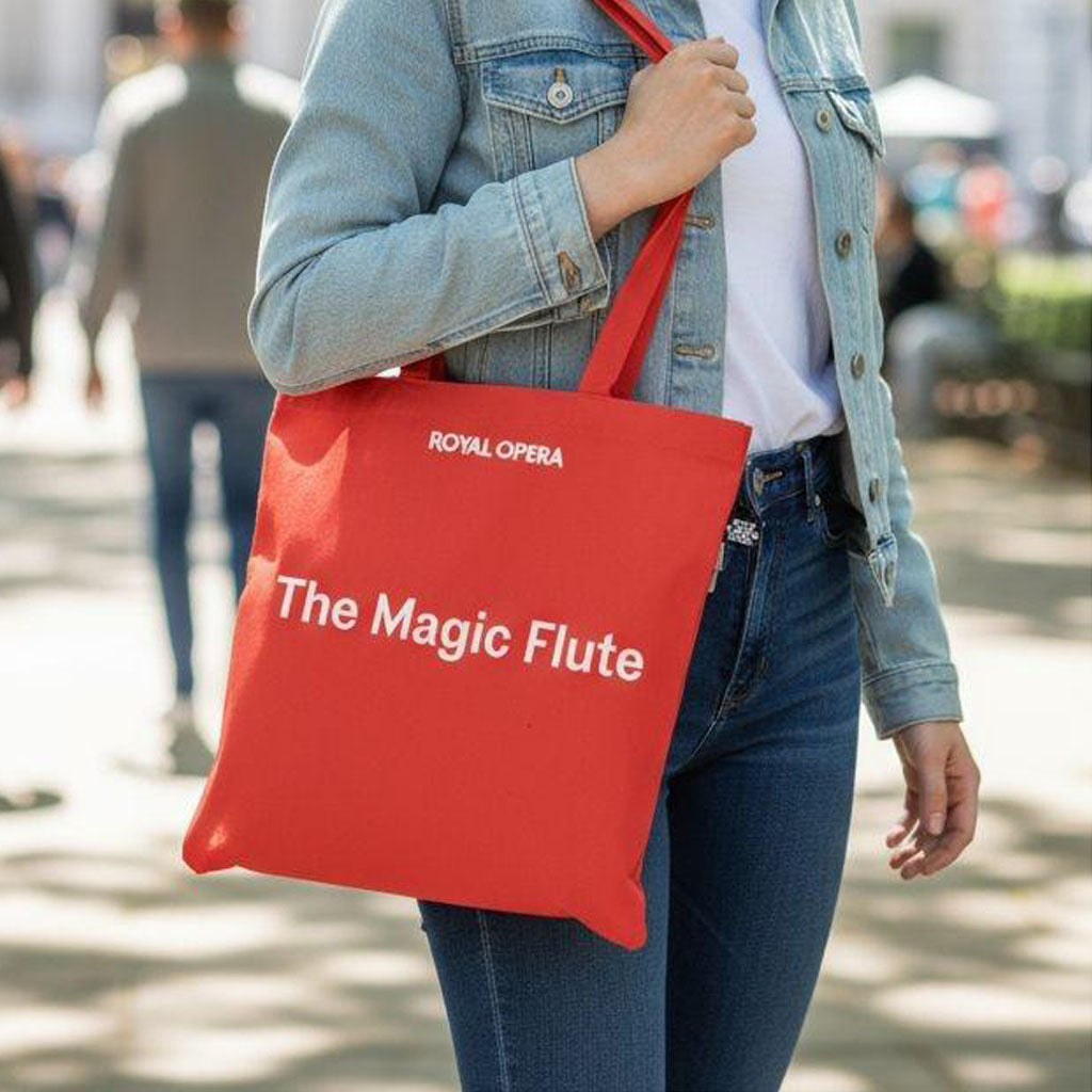 Person holding a red tote bag with &#39;Royal Opera&#39; and &#39;The Magic Flute&#39; text on a blurred street background