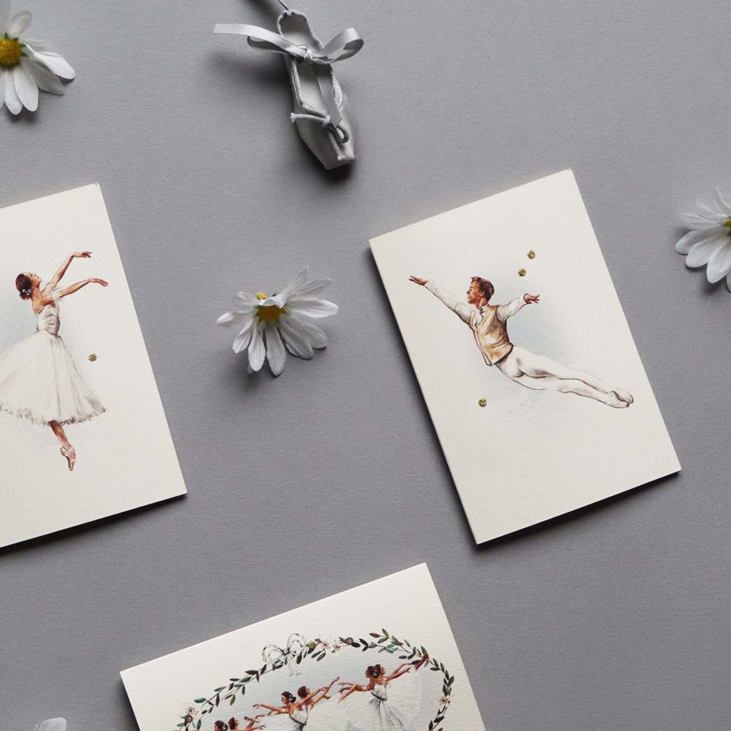 Three Giselle Greeting cards with  illustrations of dancers on a gray surface with daisies and a silver ballet shoe keyring.