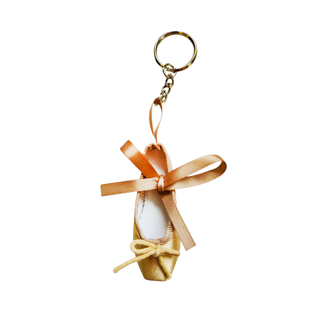 A pale gold keyring in the shape of a ballet shoe against a white background. The keyring is hanging vertically, suspended by a short metal chain with a larger ring at the top for attaching to keys. There are two gold bows, a larger one about two thirds of the way up the shoe, and a smaller one at the bottom. 