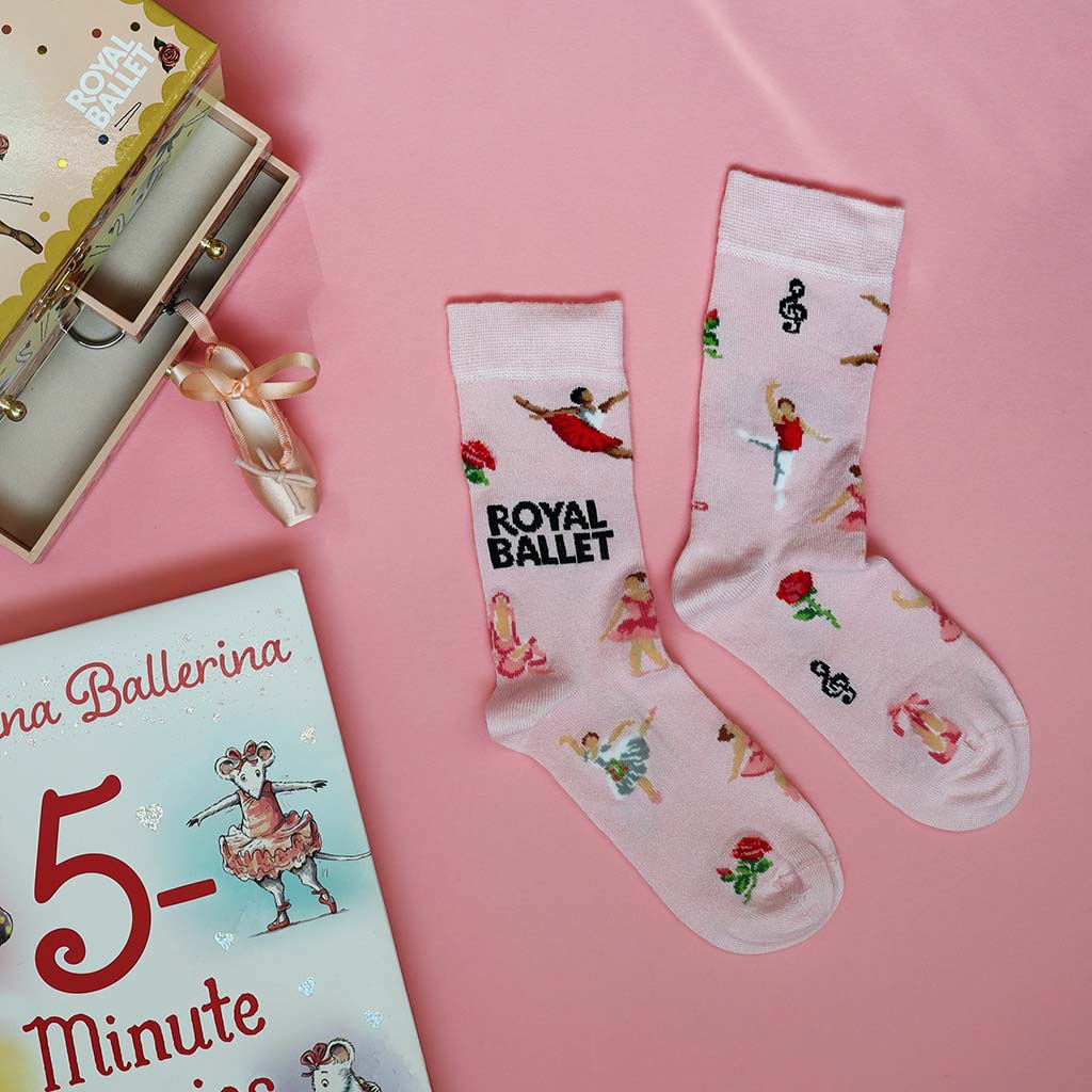 A pair of light pink socks featuring ballerina figures, musical notes, ballet shoes and flowers and the words 'ROYAL BALLET' in black text, displayed on a clean white background.