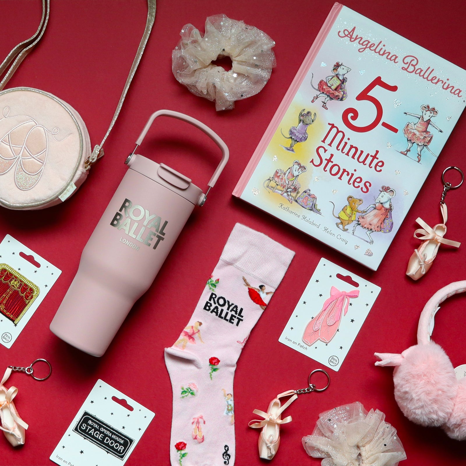 A styled flat-lay of ballet-themed merchandise on a red background, including a pale pink tumbler with 'ROYAL BALLET' text, a pink ballet sock, pink earmuffs, tulle glittery scrunchies, a pink round bag, and a children's book titled '5-Minute Stories' Angelina Ballerina.