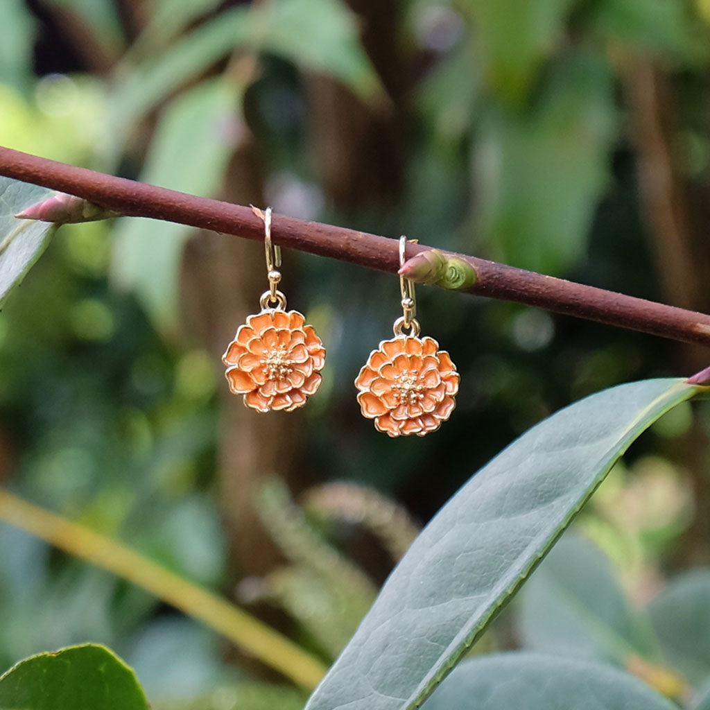 Pair of orange coloured floral earrings on a branch with green leaves in the background