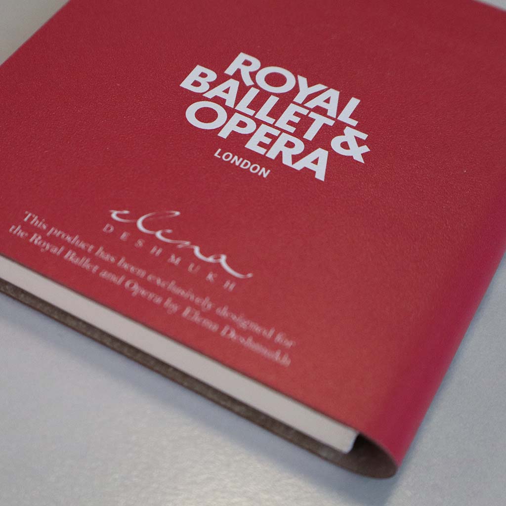 A mini notebook depicting a the auditorium at the Royal opera house  with a red velvet stage curtain, ornate gold and duck egg blue ceiling details, and an audience seated in the auditorium, shown against a plain white background.