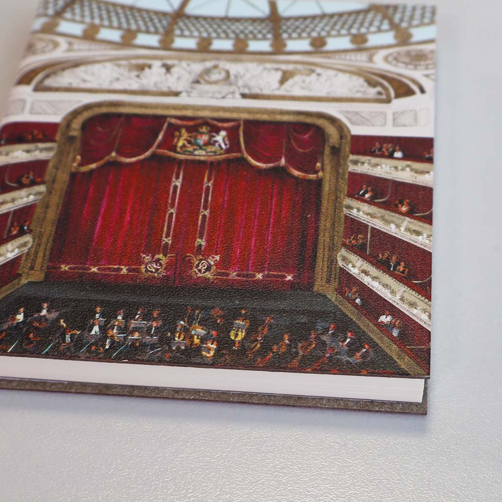 A close-up of a mini notebook depicting the auditorium at the Royal opera house with a red velvet stage curtain, ornate gold and duck egg blue ceiling details, and an audience seated in the auditorium, shown against a plain grey background.
