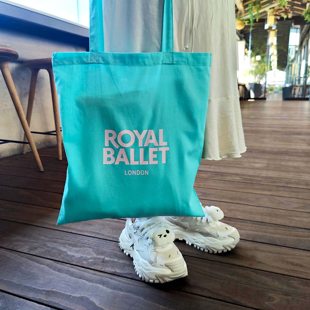 Model holding a mint green tote bag with &#39;Royal Ballet London&#39; text on the centre front standing on a terrace