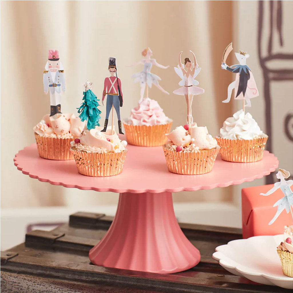 Cupcakes on a pink cake stand with decorative Nutcracker theme toppers  and gold cake cakes on a wooden surface.