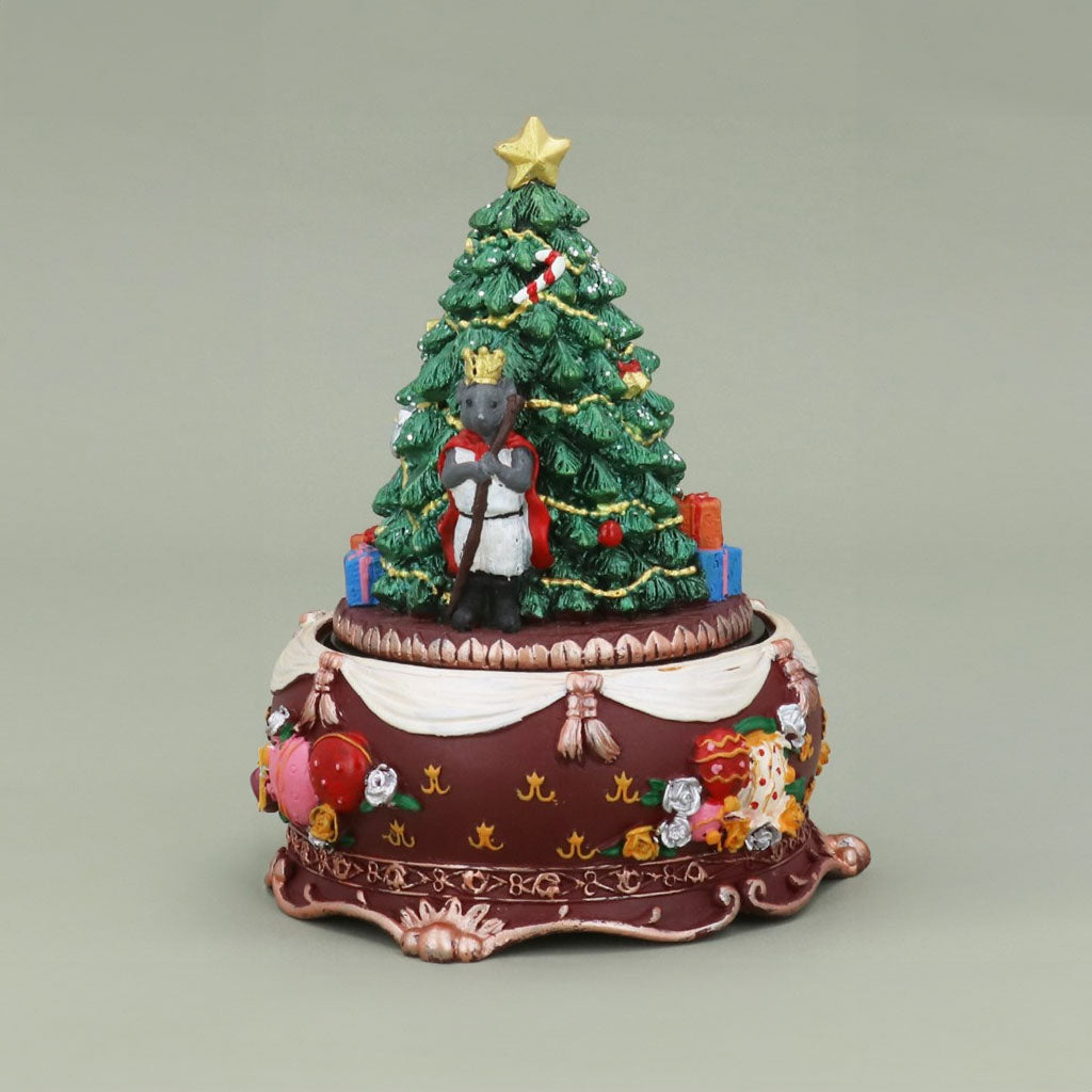 A decorative Christmas-themed figurine featuring a small mouse king standing in front of a decorated green Christmas tree with a gold star on top. The scene is set on an ornate round base with a maroon finish, white draped fabric, and colourful holiday details, photographed against a plain light green background.