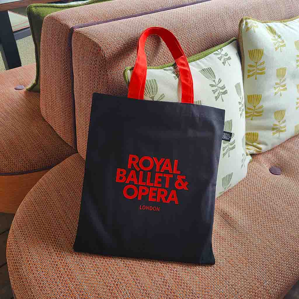 A black fabric tote bag with red handles and red 'ROYAL BALLET &amp; OPERA LONDON' text, placed on a terracotta-coloured couch with patterned cushions behind it.