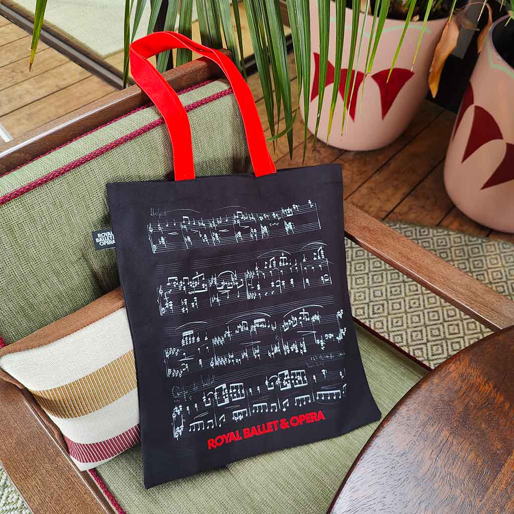 A black tote bag with red handles sits on a wooden chair in a real indoor setting. The bag has white musical notation printed across the front and red text reading 'ROYAL BALET &amp; OPERA' near the bottom.