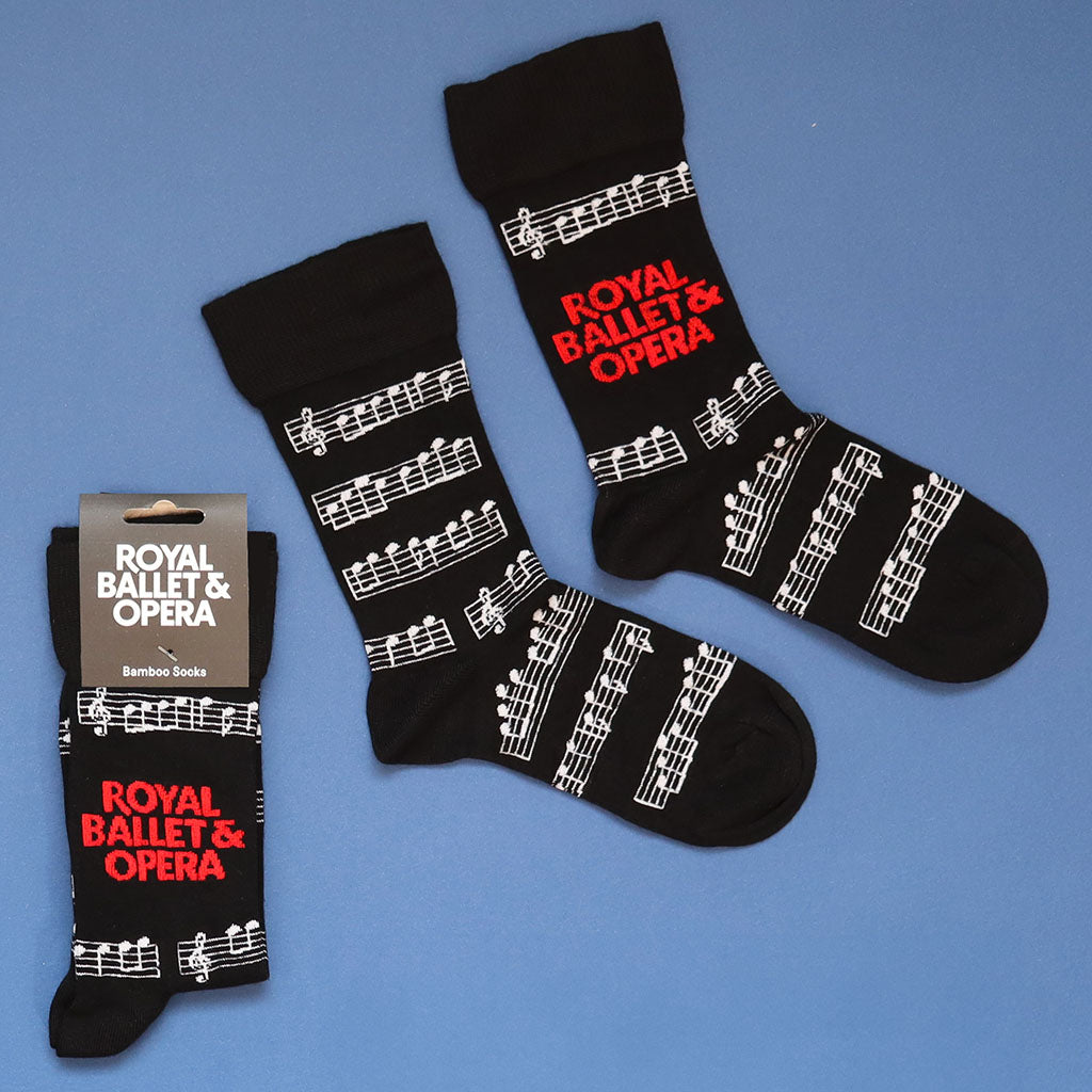 Black socks with white musical notes and staff lines, featuring 'ROYAL BALLET &amp; OPERA' in red text, shown on a blue background with one sock and its retail packaging.