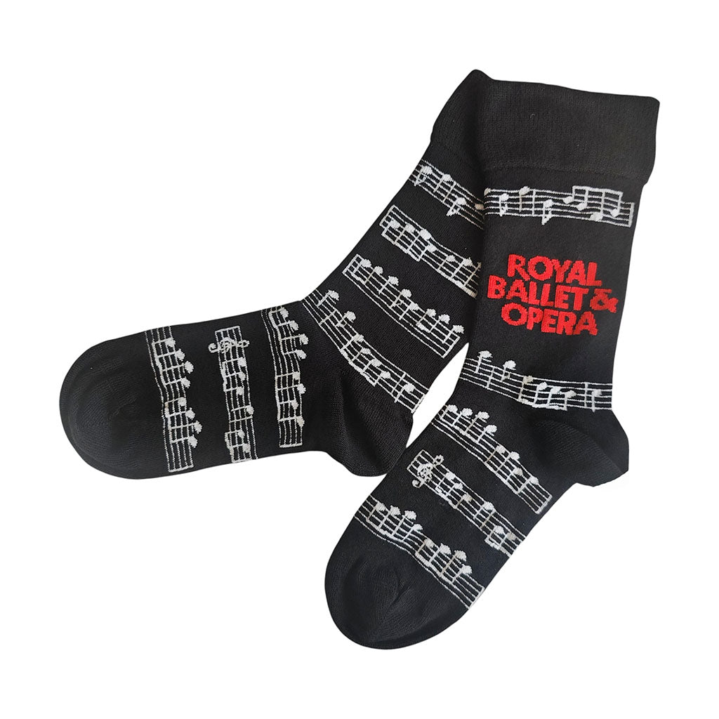 A pair of black socks featuring white musical staff and notes printed across the body, with 'ROYAL BALLET &amp; OPERA' written in red text on one sock, shown against a plain white background.
