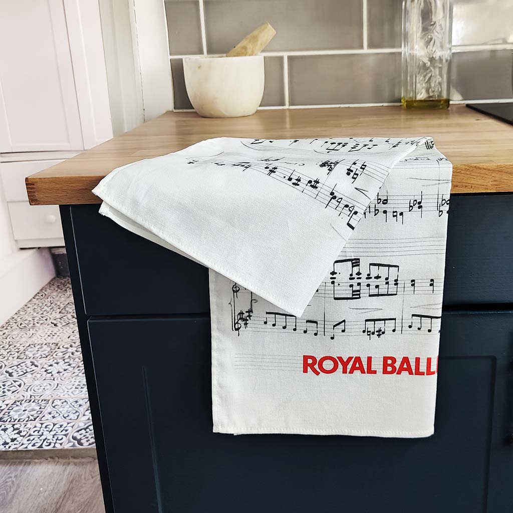 A white  kitchen tea towel printed with black musical sheet music and the red text 'ROYAL BALLET &amp; OPERA' hanging over a dark blue kitchen cabinet.