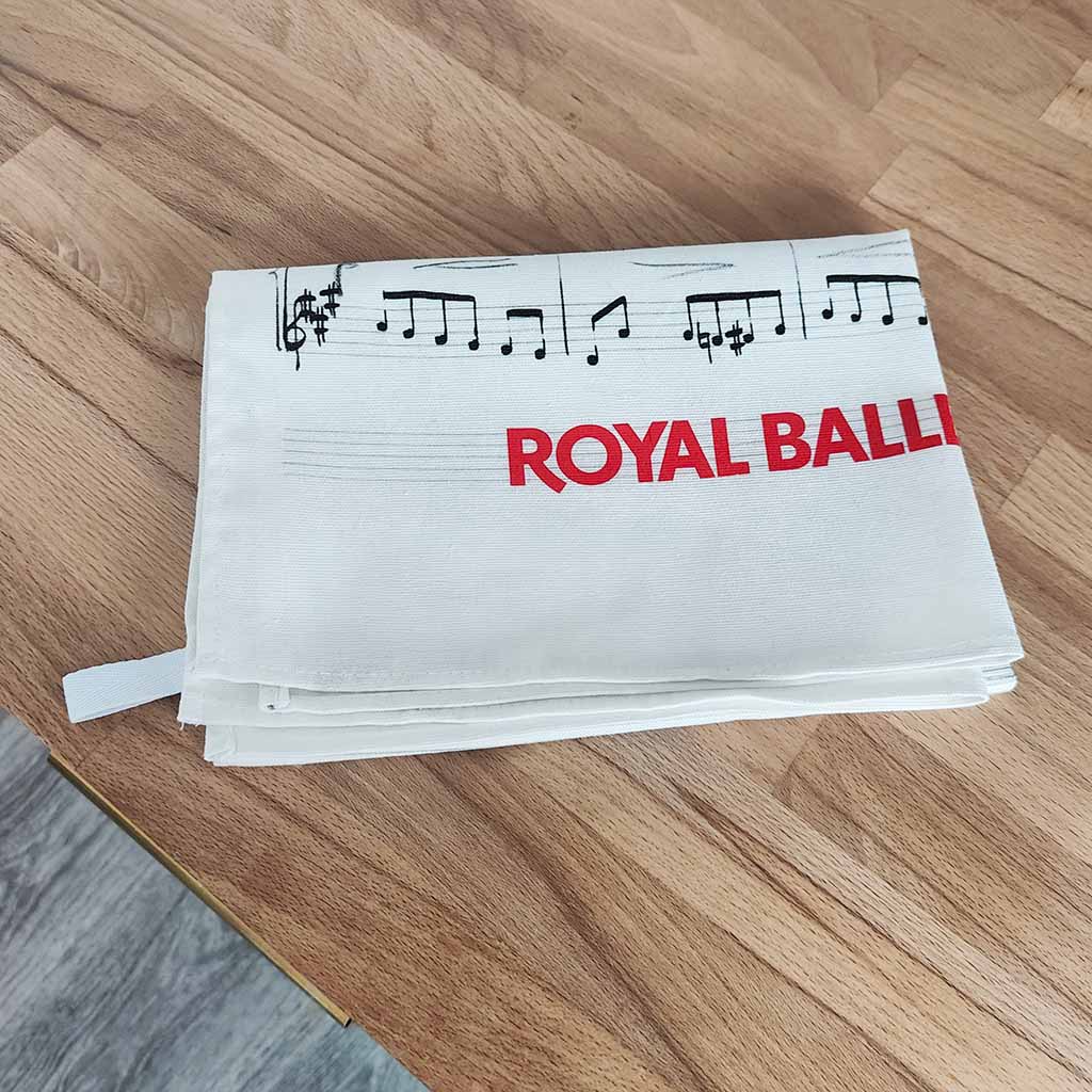 A folded white Royal Ballet and Opera Tea Towel featuring a music manuscript  design  resting on a light brown wooden worktop.