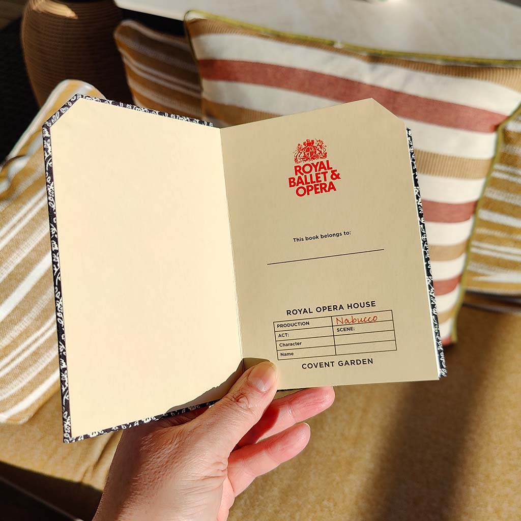 Hand holding a Royal Ballet &amp; Opera notebook with a striped chair in the background