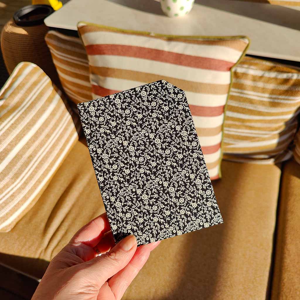 Hand holding a black and cream fabric notebook with white floral pattern in front of striped cushions.