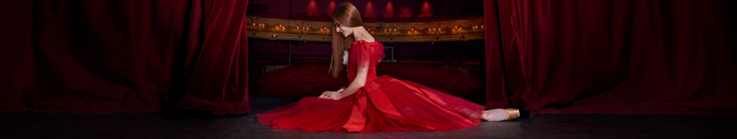 Marianela Nunez in a dramatic red gown sits on a dark stage floor in front of red curtains and a gold-trimmed backdrop.