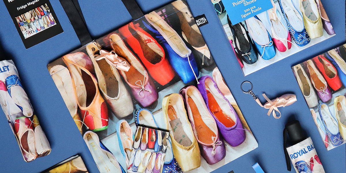 A styled flat-lay of ballet shoe-themed merchandise on a blue surface, including a large printed bag with colourful ballet  shoe tote bag, jigsaw, magnet , a small keyring, and an umbrella labeled Royal Ballet.