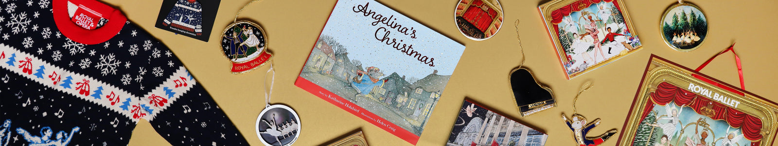 A styled flat-lay of various Christmas-themed merchandise on a beige background, including a navy blue knit sweater with white snowflakes, a 'Angela's Christmas' book, a red Santa hat, and several round and rectangular ornaments.