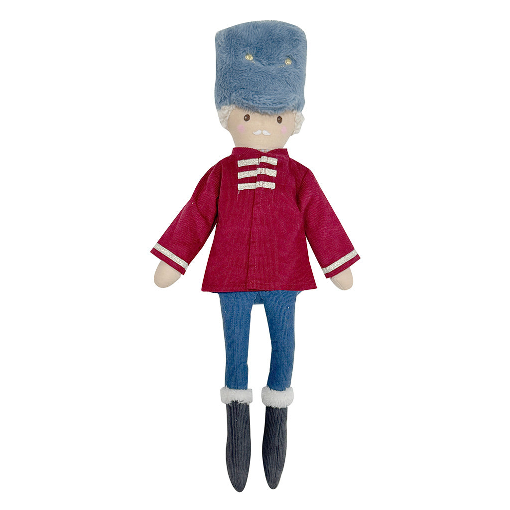 A plush doll dressed as a nutcracker soldier from the ballet The Nutcracker, wearing a blue fur-look hat, a magenta-red jacket with gold trim, blue pants, and dark gray boots, isolated on a white background.