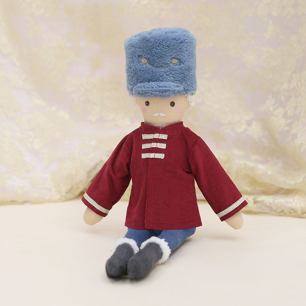 A plush doll seated against a soft cream lace fabric backdrop. The doll wears a blue fuzzy hat, a burgundy-red jacket with gold trim, blue pants, and dark gray shoes.