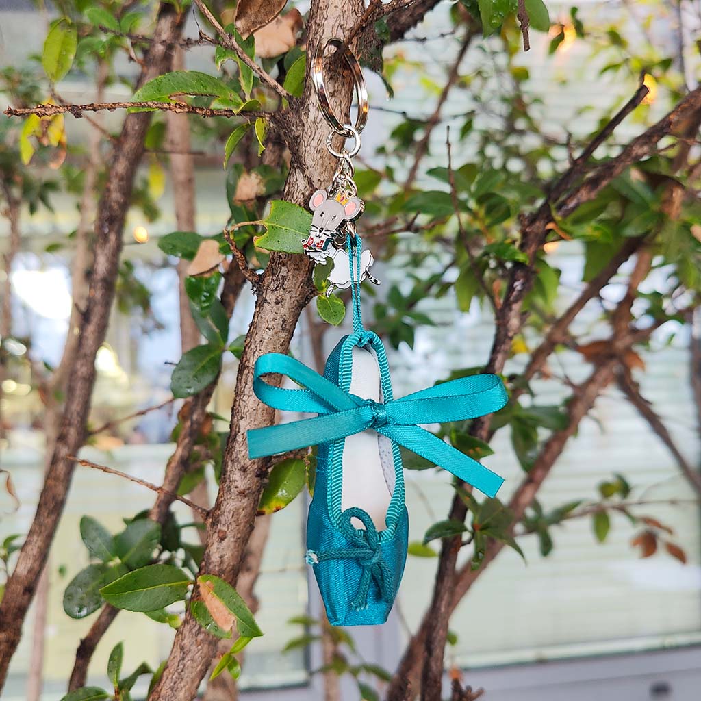 A miniature ballet shoe keyring  hanging from a tree branch outdoors. The shoe is a teal turquoise with a ribbon tied around it, and small decorative charms are attached at the top hanging on a branch