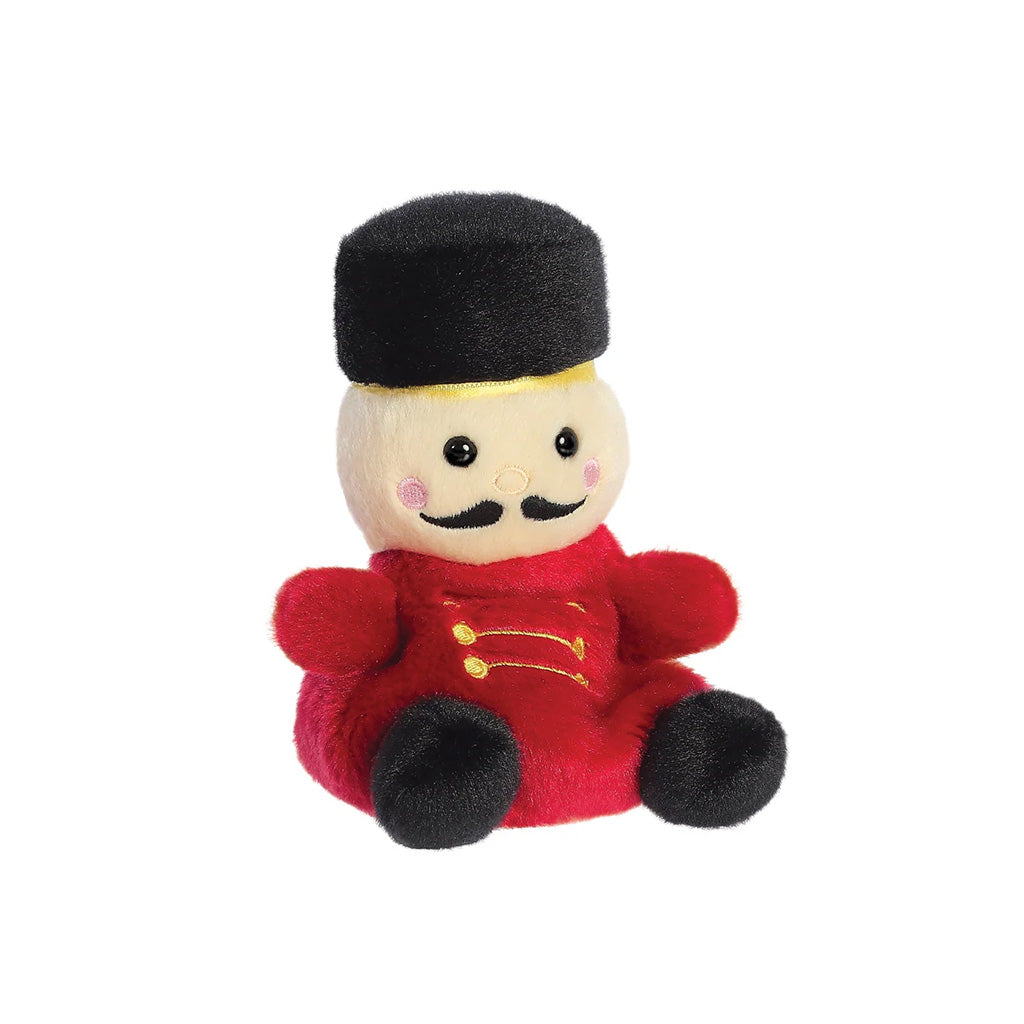 A small plush toy dressed like a nutcracker, with a cream face and rosy cheeks, black hat , moustache and  shoes, red jacket with gold button details, isolated on a white background.