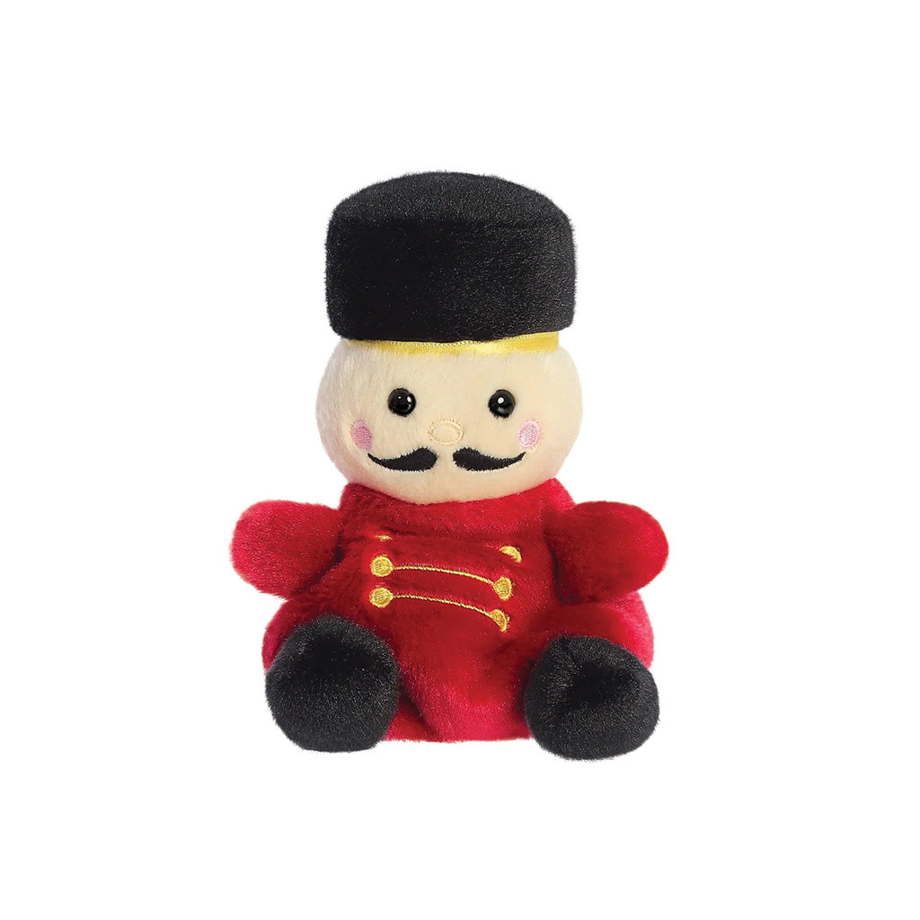 A small plush toy dressed like a nutcracker, with a cream face and rosy cheeks, black hat , moustache and  shoes, red jacket with gold button details, isolated on a white background.