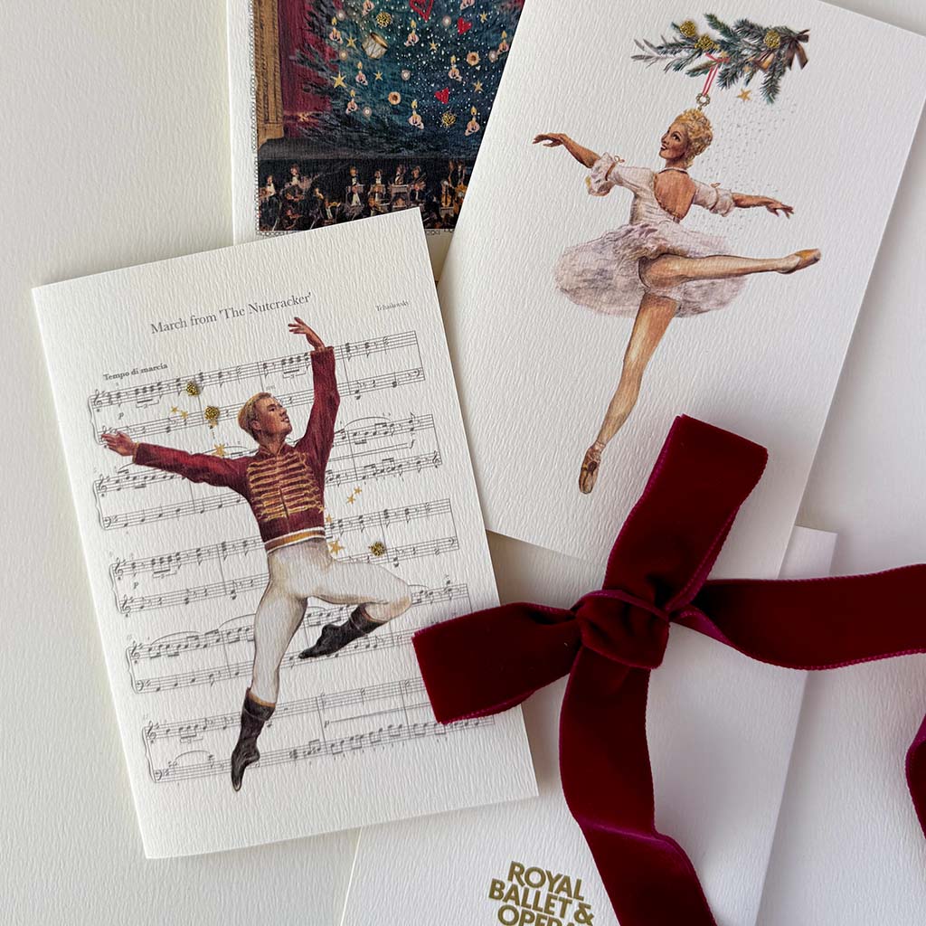 A styled flat-lay of ballet-themed greeting cards, featuring a white card with a Nutcracker dancer illustration over sheet music, another ballet dancer card, and a dark red velvet ribbon bow. A gold logo at the bottom reads Royal Ballet &amp; Opera.
