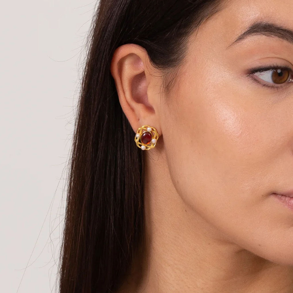 A close-up of a woman's ear wearing a gold stud earring with a circular design and a central red gemstone and white pearls.