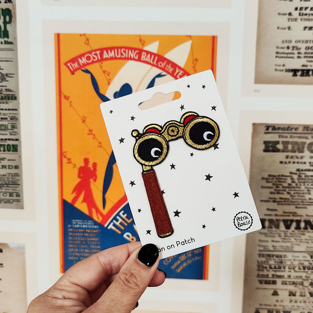 Hand holding an iron on patch of opera glasses on a card with vintage theatre posters background