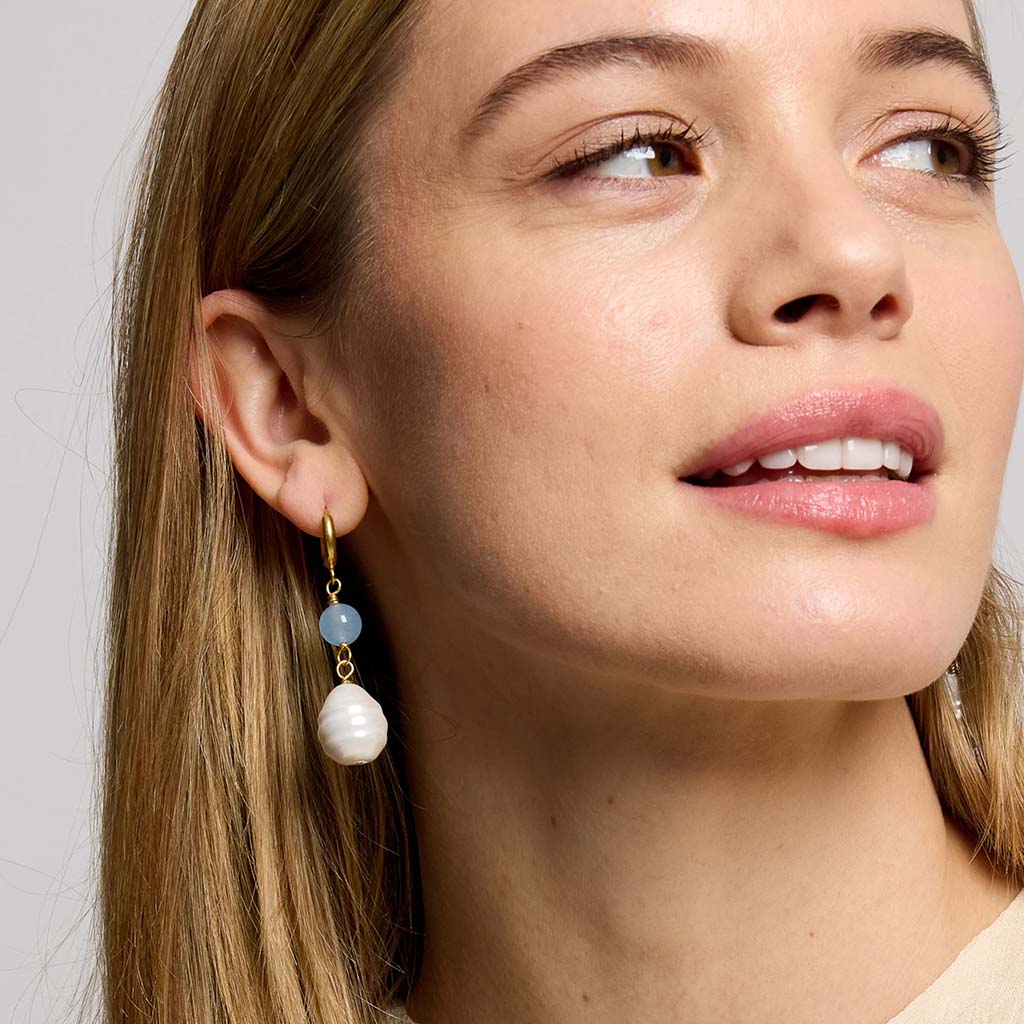 A close-up of a woman wearing a gold drop earring with a light blue stone, a white baroque pearl, and a small white bead.