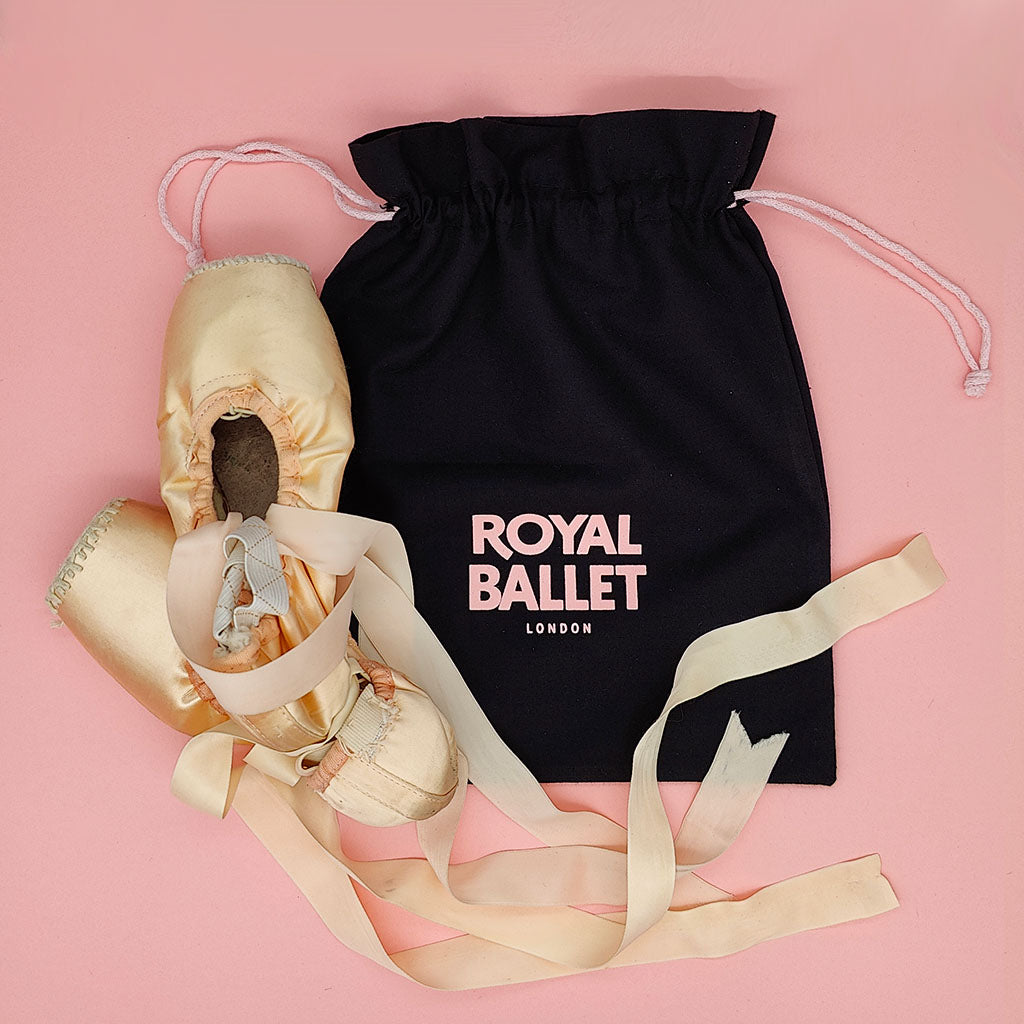 Ballet shoes with ribbons and a black drawstring bag labeled 'Royal Ballet London' on a pink background.