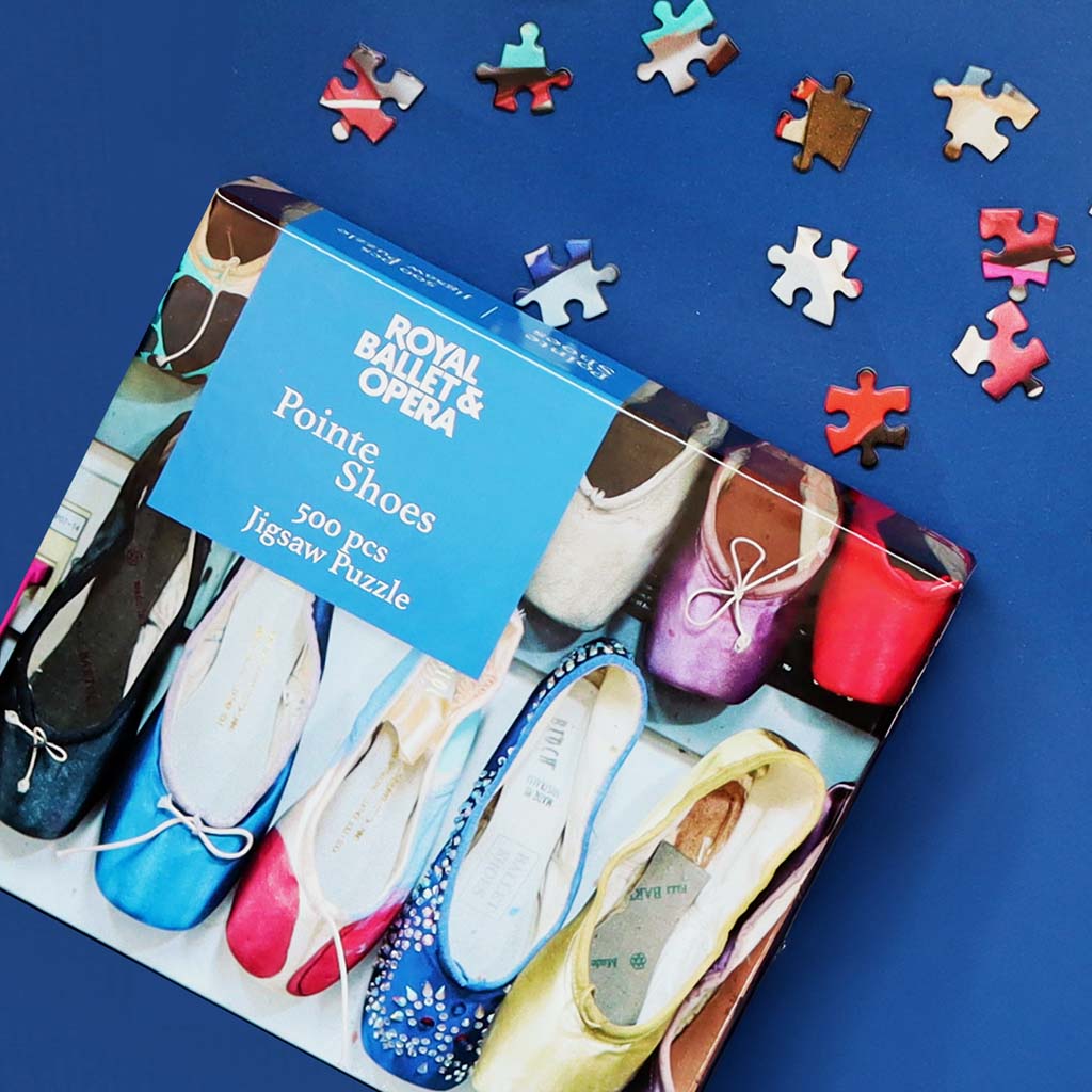 A boxed jigsaw puzzle titled 'Royal Ballet &amp; Opera - Pointe Shoes' lies on a blue background with several loose puzzle pieces scattered around it. The box shows several ballet shoes in different colours, and the puzzle itself appears to be a 500-piece puzzle.