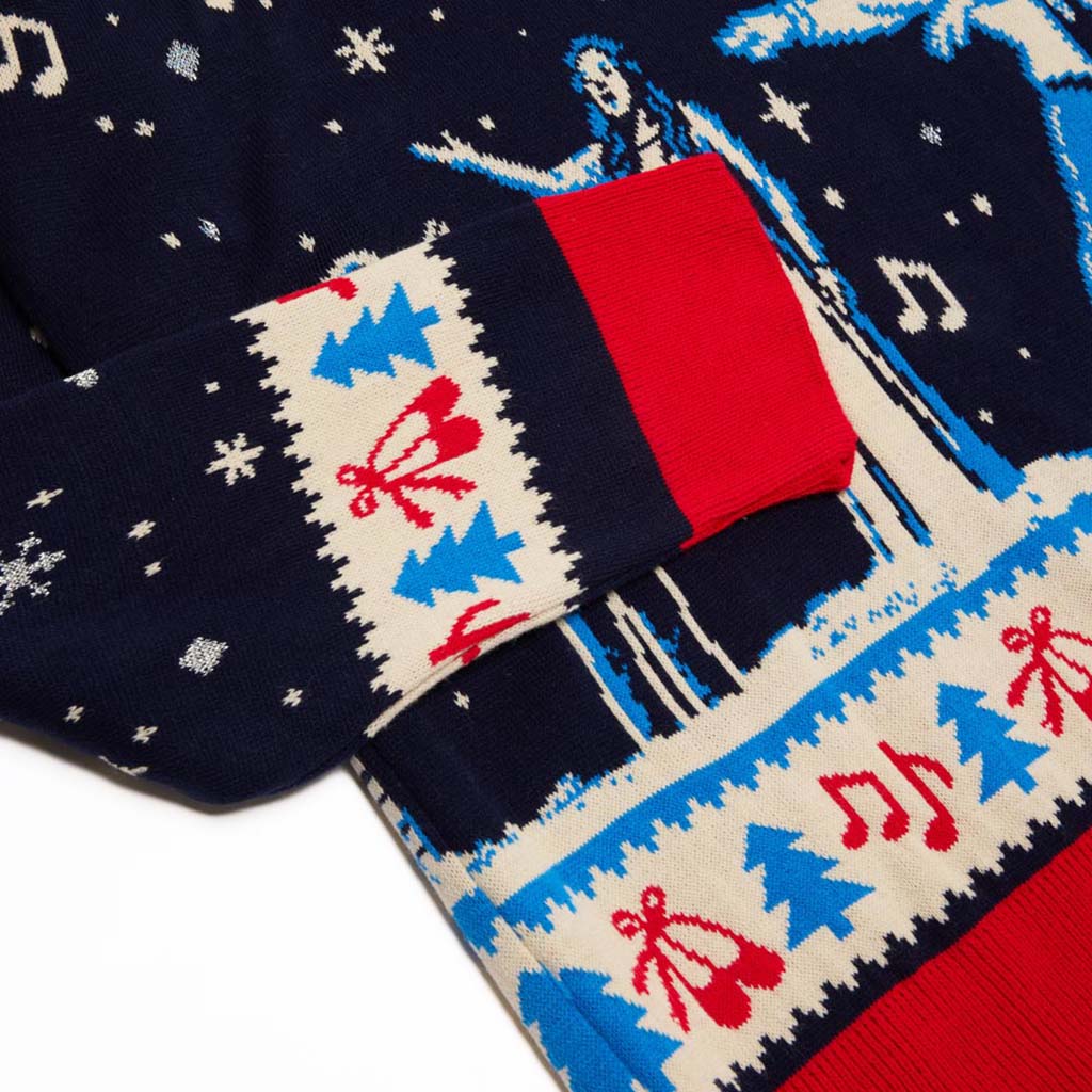 Close-up of a navy blue Christmas sweater with a festive knit pattern featuring red ballet shoes, blue Christmas trees, white snowflakes, and a red cuff.