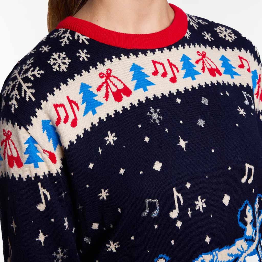 Close-up of a navy blue Christmas sweater with a red crew neck, featuring a cream band with blue Christmas trees, red ballet shoes, and red musical notes, plus scattered white and blue snowflake and star motifs on a light grey background