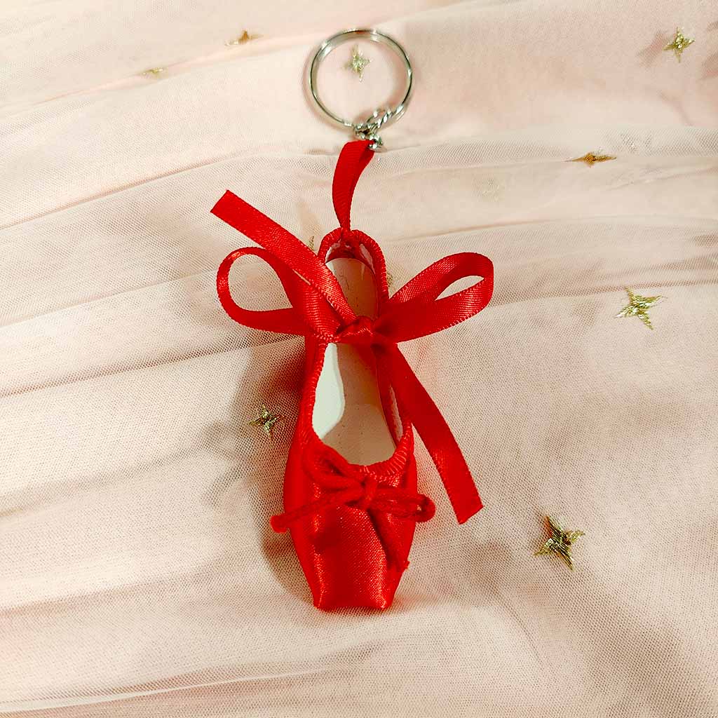 A Red Ballet Shoe Keyring lying on a pink tulle and lace background. On the lace there are embroidered stars with 4 points. 