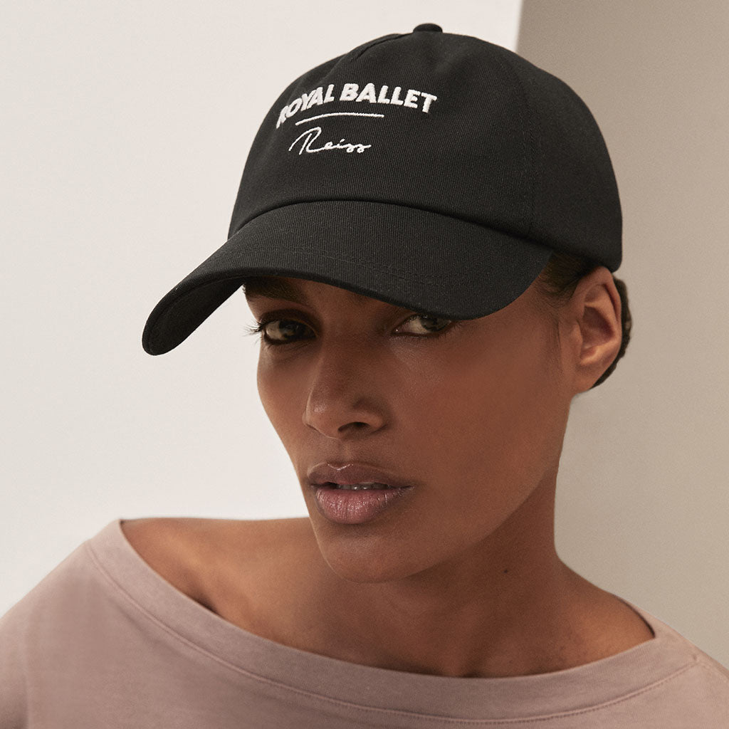 Person wearing a black cap with &#39;Royal Ballet Reiss&#39; text on a neutral background