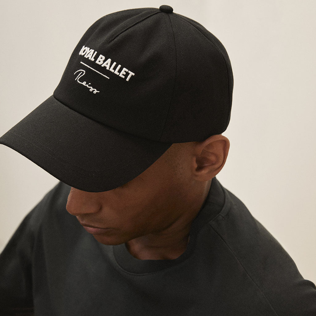 Person wearing a black cap with &#39;Royal Ballet Reiss&#39; logo on a neutral background