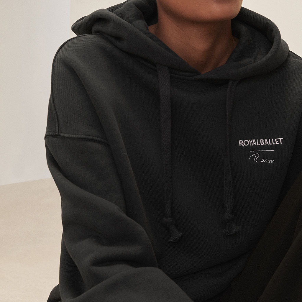 person wearing a black hoodie with a brand logo Royal Ballet Reiss, posing against a neutral background